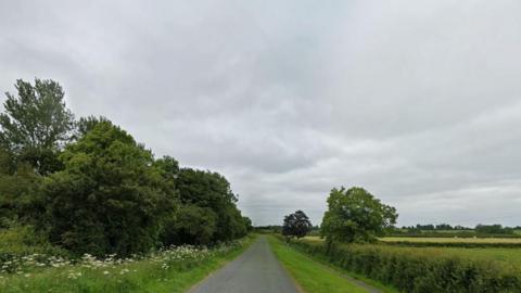A countryside road. Grass verges with green bushes and trees line the road. The sky is overcast. 