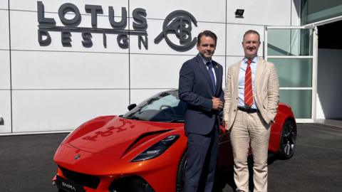 Two men standing in front of a sleek, red Lotus Emira car at the Lotus plant.
