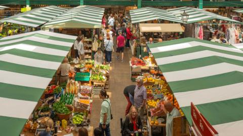 Large, stripey green roof covers stand above the stalls where food, groceries  and clothes can be seen with shoppers walking around them.
