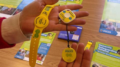 A woman is holding the three devices that are part of the scheme. All three are yellow. One is a wristband, one is a square tracking tile, and the last is a round disk on a strap which acts as a hangtag. 