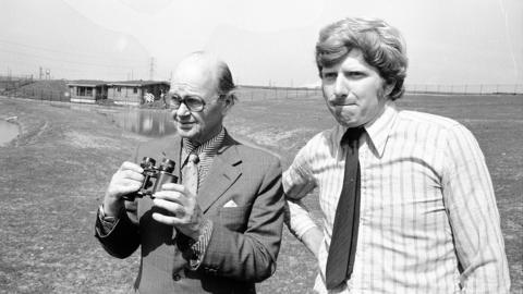 A black and white photograph of Sir Peter Scott visiting the wetlands on 20 May 1975. He is wearing a suit and tie and holding a pair of binoculars. He has glasses. Curator John Secrett is standing next to him and pursing his lips as he looks into the camera. He is a wearing a white striped shirt with a tie. The pond and the visitor centre building are behind them. 