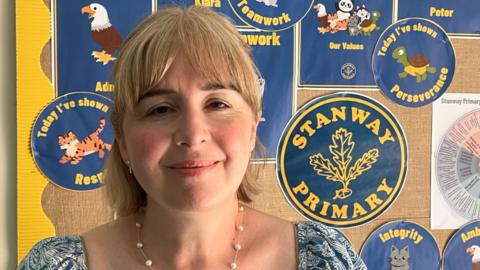 Rebecca Loader stands in front of a noticeboard with various pieces of paper on display, including a navy blue circle with yellow writing which says Stanway Primary, with a trio of leaves in the middle. Rebecca has blonde hair and is wearing a blue and white floral top with a necklace