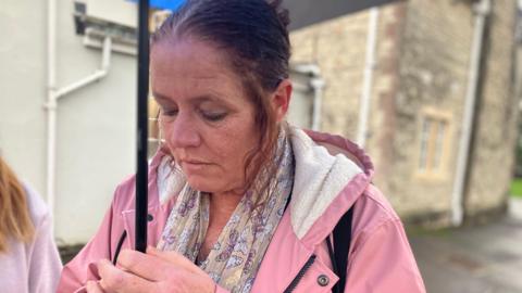 Martine, Mr Cole's mother, is standing under an umbrella looking down on the ground. She is wearing a pink jacket.