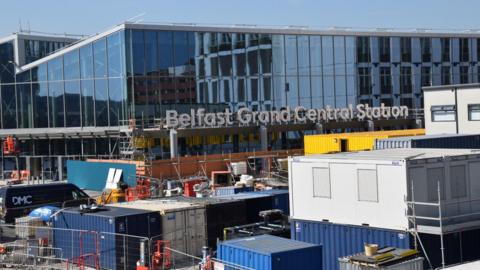 Belfast Grand Central Station is a large building. The roof is zig-zag shaped. Glass windows cover the entire building. There are numerous cargo boxes and fences around the building.