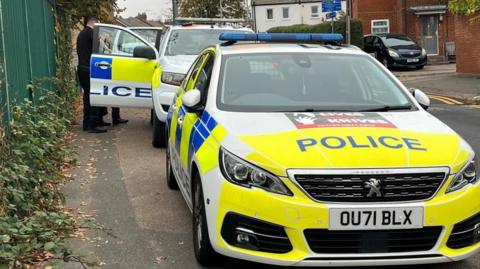 A number of police cars in a street, with police officers, dressed in black by a car. There are homes in the distance and a car parked on a drive.