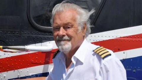 Elfan Dyfed ap Rees wearing a white collared shirt with gold and black striped epaulettes on the shoulders. He has white hair and a white goatee-style beard. He is smiling at the camera, standing beside an aircraft painted with blue, white and red stripes.