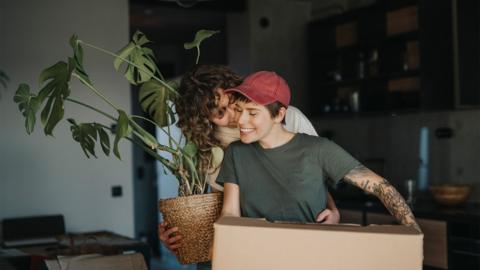 
Smiling woman holding houseplant kissing girlfriend carrying cardboard box moving house - stock image