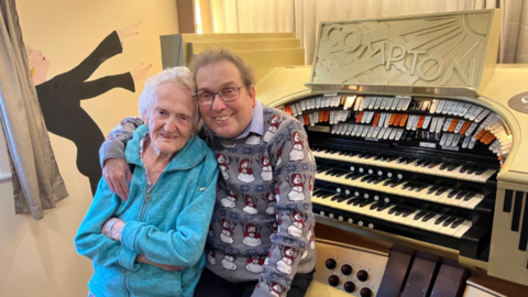 Edna and Simon, with his arm around her, sitting in front of the organ in a small room