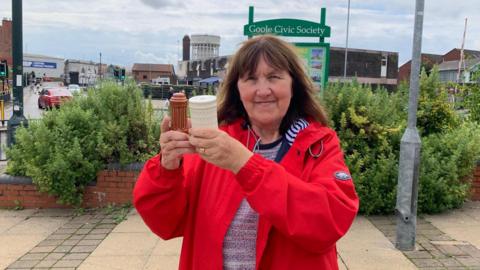 Margaret Hicks-Clarke wearing a red coat and with brown shoulder-length hair holds up the ceramic salt and pepper pots. They resemble the town's twin water towers which are behind her in the picture. 