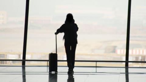 The silhouette of a person with a suitcase standing at a big glass window at an airport 