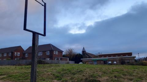 The former site of the Eagle Pub. It is an empty lot covered in green grass. A building and a church stand in the background. A sign post for the former pub stands in front of the grass, but the sign itself is missing.