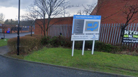A blue sign reading Whitworth Road Industrial Estate displays a map with the units on the site. Behind it, there is a grey metal fence and a brown brick building.