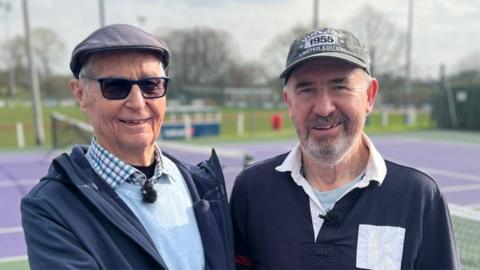 Head and shoulders image of David Pearson and Andrew Lockwood standing outside at Garstang Tennis Club by a purple court. David is wearing sunglasses, a flat cap, and a blue checked shirt under a pale blue jumper and dark blue coat. Andrew is wearing a baseball hat and navy and white rugby shirt. He has a short greying beard