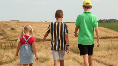 Three children, a little girl, a boy and an older boy, walk along dry grass near the coast. They have their backs to the camera. It's a summer's day.