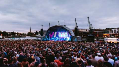A crowd of people watch a musical act on a lit up stage on Bristol harbourside with the dock's cranes in the background