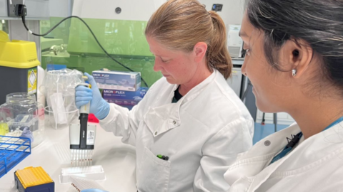 A woman in a lab coat, watched by another woman in a lab coat, uses lab equipment