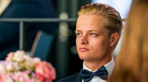 Marius Borg Høiby, who has gelled back blonde hair and a gold hoop earring, wears a tuxedo with a black bow tie as he looks into the distance at a formal event