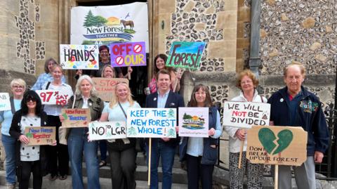 Group of people stand outside Hampshire council chamber with signs saying "don't break up our forest", "Keep the New Forest whole" and "we will not be divided"