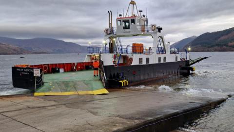 The Maid of Glencoul is a small ferry painted black and white with a green car desk. A ferry worker in bright orange overalls is standing on the car deck.