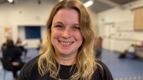 Ellie Short has blonde wavy long hair and is smiling at the camera. She is wearing a black T-shirt with a Herts Inclusive Theatre badge.