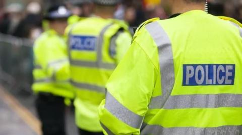 A police officer in a yellow hi-vis jacket. The word "police" is on the back. There are two other officers in the background in the same jackets with black police hats on