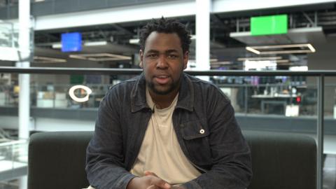 A man in a dark denim jacket and a beige t shirt is sat down looking straight at the camera. He has spiky afro hair and there is a small mic poking out of his t-shirt. In the background there are blurry tv screens with various different output from the BBC. There is also a glass wall roughly a waist height and two coloured squares, one blue and one green.