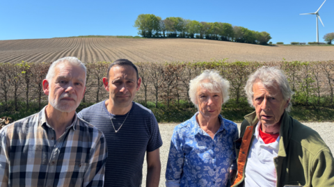 Three men and a woman stand in front of a field. A trimmed hedge separates the road from the field. Green trees are in the distance. A large wind turbine is located behind the field. The sky is blue.