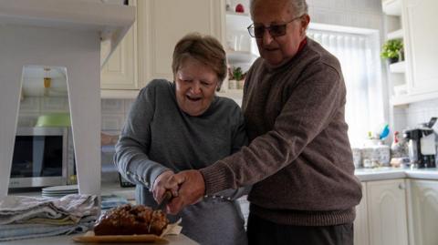 Doreen and Michael Binder stood at their kitchen counter holding a kitchen knife together and cutting a cake