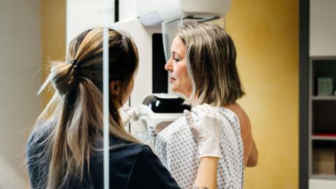Stock image of a grey-haired woman with a nurse in a blue gown. The woman is wearing a white medical gown and standing sideways by medical equipment. She is being helped by the nurse who is wearing white gloves. The woman looks nervous. You can only see the back of the nurse's head.