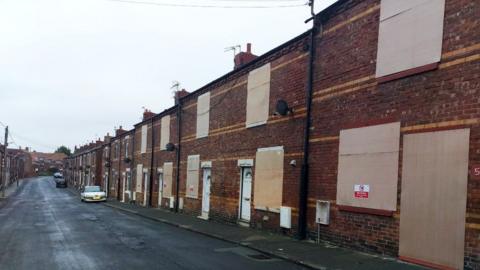 A row of red-brick houses with almost every window and door boarded up