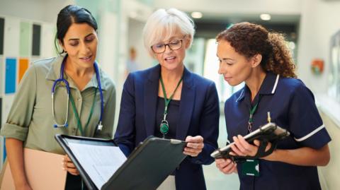 A woman middle-aged woman wearing a shirt with dark hair and a stethoscope around her neck, an older woman in a blazer holding a file, and a younger woman in a nurse's outfit holding a tablet computer, all stand next to each other in a hospital ward