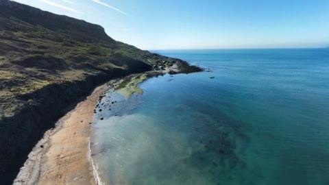 Rugged curved cliffs with sandby beach and green/turquoise sea - it is a calm sunny day.