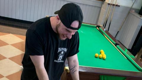 Tyran in a room with a pool table. He is smiling and looking own. He is wearing a black t-shirt and black cap. He has a tattoo on his left arm.