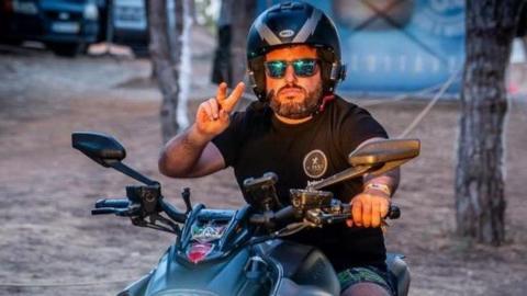 A man doing a peace sign while on a motorbike and wearing a helmet. It is a warm looking evening and he wears a black T-shirt and shorts.