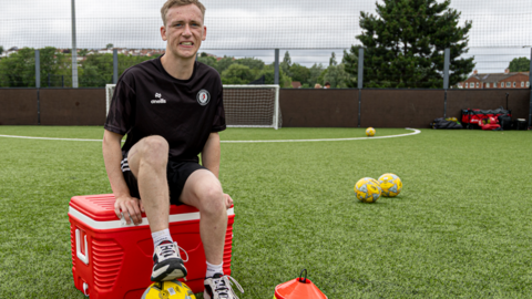 A man is sitting on a red box, and has his foot placed on top of a football. Behind him is a small goal, and various bits of football training equipment.