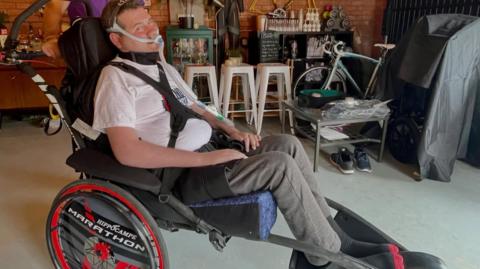 Sam Perkins sat in his race chair. He is wearing a white t-shirt and joggers.