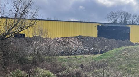 The image shows a yellow warehouse in the distance with a mound of brown industrial waste in front. Rubbish can also be seen piled up inside the building.