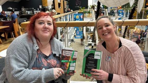 Carys Wiggans, on the left, with red hair, wearing a grey cardigan and a blue top with roses on it. She has a ring on her middle finger and her ring finger. She is Holding a book, titled The Great Theatre Rescue, which is wrapped in green ribbon and has a book fairy sticker on it. Kate Pearson, on the right, wearing a pink jumper, her blonde hair is pulled back. She is wearing a necklace, the pendent is square and is multi-coloured with white, pink and blue. She is wearing a ring on her ring finger. She is holding a green book, with pink ribbon on it and a book fairy sticker, with a bookmark which says 'Free Book' sticking out of the top. 