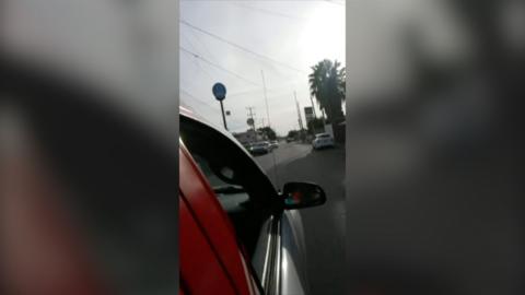The right-hand front door and wing mirror of a red car seen from behind as it travels down a road Culiacán, Mexico.