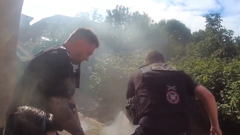 Three police officers are seen bending over a suspect on a paved area outside a house. The suspect lying on the ground cannot be seen. A cloud of thin smoke is rising around the officers.