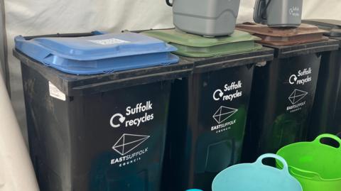 Four black bins with different coloured lids are lined up inside a tent during an event. Small grey food caddie bins sit on top of the bins.