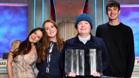 A group of four people stand side by side - left to right, a woman with long dark hair and a pale flowery dress who has her head on the shoulder of a woman with long red hair and wearing a blue top, her arm around the other woman, a person in a blue beanie and blue top holding a silver award in the shape of an open book, and a man with short dark hair and a dark jumper. 