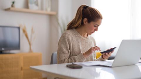 Woman sits at a desk in a home with a phone and a laptop in front of her and a calculator in her hand.