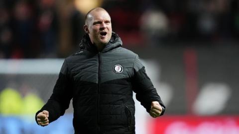 Liam Manning wearing a black and grey puffer jacket with a Bristol City badge on the chest. He is pumping his fists at his sides.