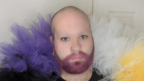Phil Harper. He has pink facial hair and purple, white and yellow ruffles material around his head. He is standing in front of a plain white backdrop and looking directly into the camera.