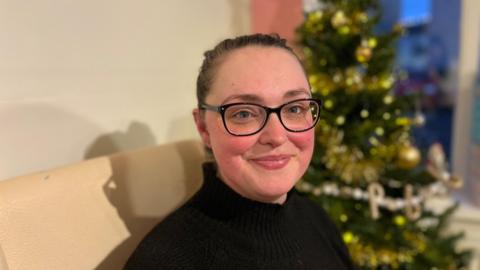 Lisa Myatt inside the Communitea hub in Wigton. She has dark hair tied up in a pony tail and is wearing glasses. She is smiling at the camera and there is a Christmas tree behind her.