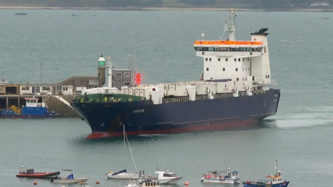 The freight ship Arrow, with a blue hull and white upper deck, sailing in to port with small boats in the foreground and a breakwater in the background