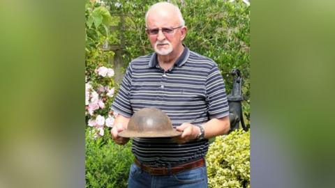 Dennis O'Callaghan, with white hair, beard and glasses, wearing a blue striped short-sleeved shirt while stood in a garden. He is holding a World War One British helmet, which was refurbished in the late 1930s and used during World War Two.