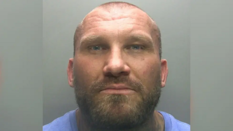 Mugshot of Derek Heggie, who is bald with a broad build and wearing a blue top. He has a short beard and moustache flecked with lighter coloured hairs.