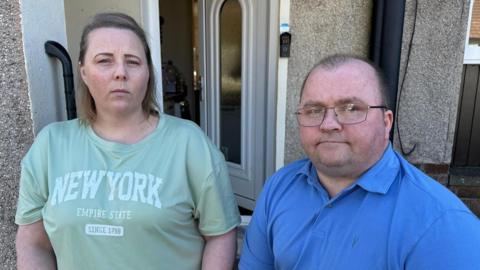 Chris and Amy Masterson standing outside their front door. Amy is wearing a green t-shirt and Chris is wearing a blue poloshirt and glasses.